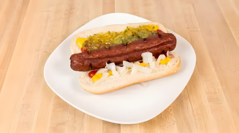 Two hot dogs with mustard, relish, and onions on a white plate on a wooden table.