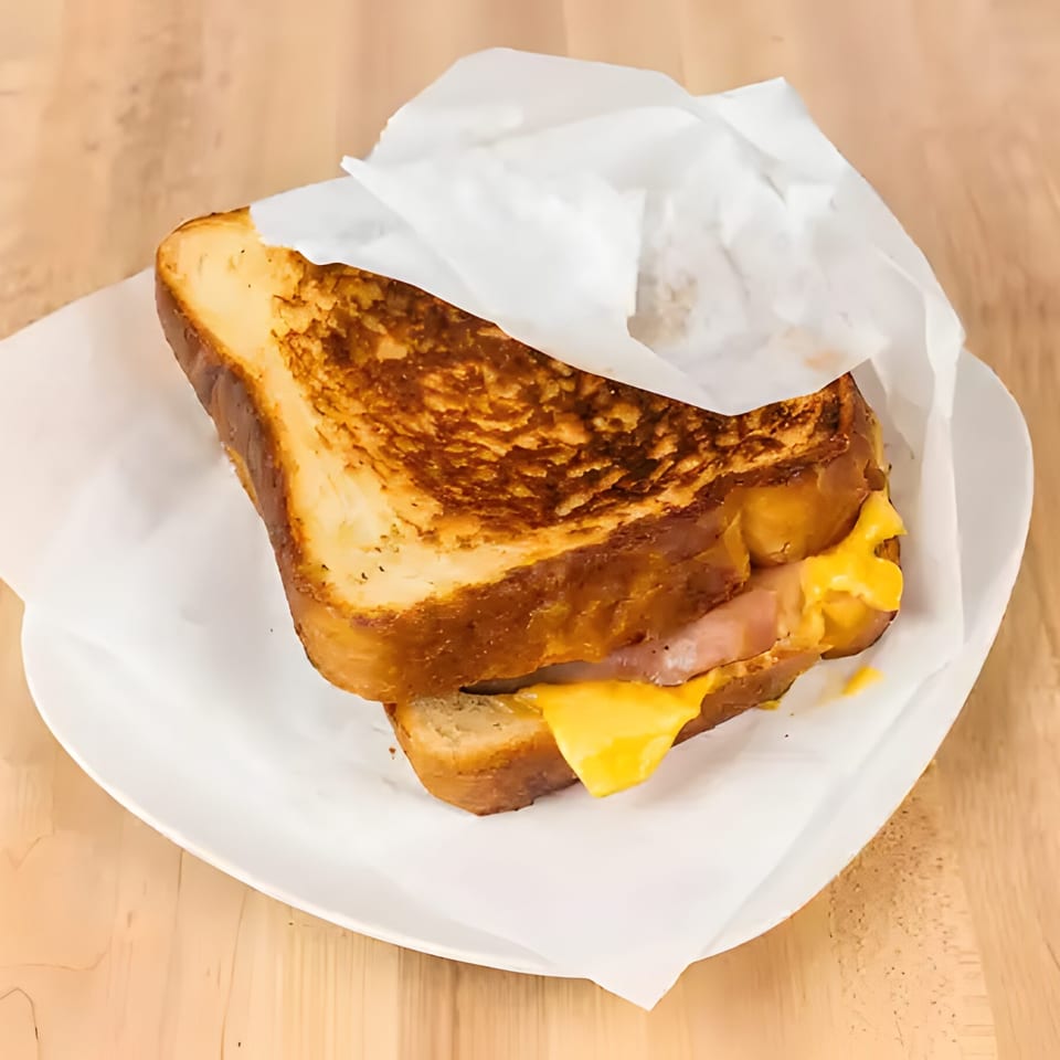Grilled cheese sandwich with melted yellow cheese and ham, wrapped in paper on a white plate.