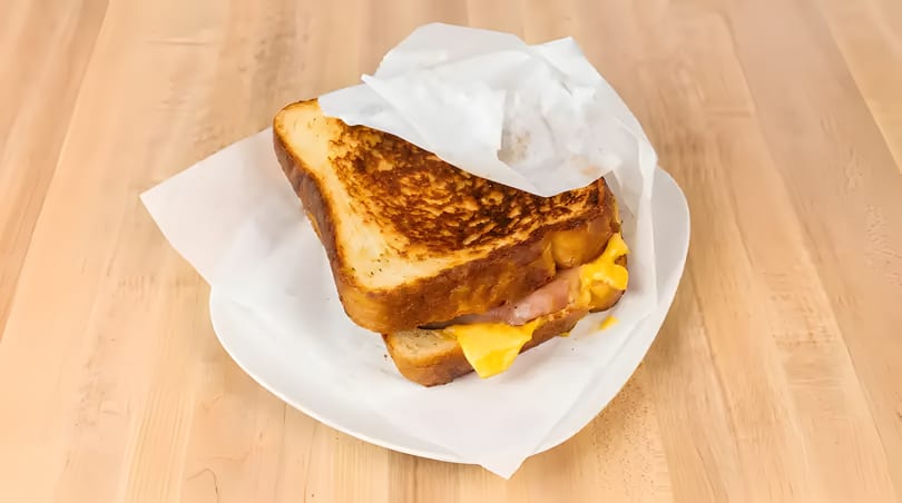 Grilled cheese sandwich with melted yellow cheese and ham, wrapped in paper on a white plate.